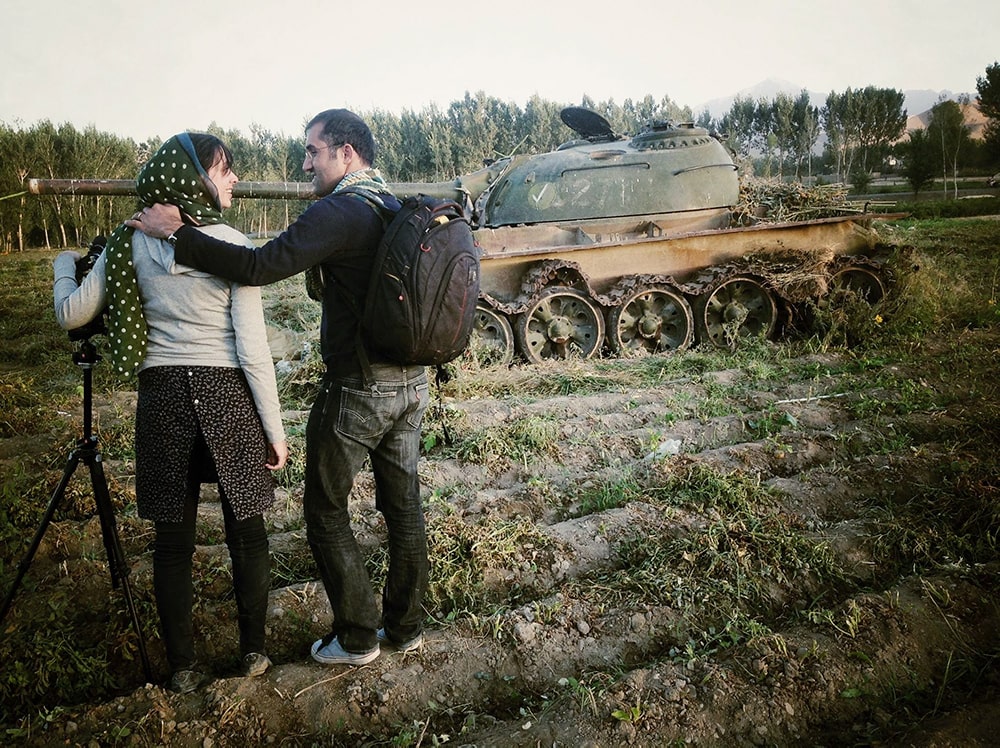 Elizabeth Mirzaei is a white woman wearing a polka dotted headscarf and holding a film camera. She is standing with partner, Gulistan Mirzaei who has his arm on her shoulder. They are in Afghanistan, in 2013, standing in front of a discarded army tank. Image courtesy of the authors.