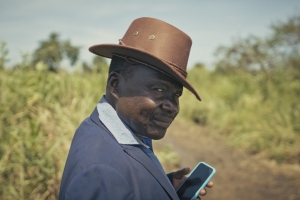 Photo courtesy of CPH:DOX. Still from 'Theatre of Violence.' A person with dark skin, wearing a brown rimmed hat and a blue coat, holding a cellphone, stands in a green field with a dirt path.