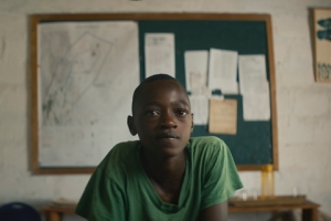 A teenaged boy wearing a green shirt is leaning forward and looking at the camera.