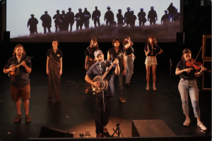 A man stands strumming a banjo in the middle of a stage, singing into a microphone. Other musicians and players surround him while “The Edge of Nature” plays on the screen behind them all.