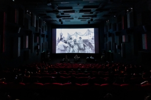A large, dark theater screening “This Is Not a Film by Deng Nan-guang” with an experimental performance using drums and other instruments on the stage below the screen. 