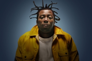 Photograph of a Black man with a yellow jacket, leaning into the camera.