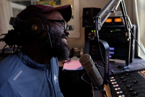 A black man sits in front of a microphone, smiling.