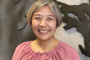 Headshot of Asako Fujioka. An east-Asian woman smiles at the camera with a concrete wall with black decorations behind her. She has straight graying hair and is wearing a loose dark pink blouse.
