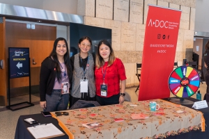Three Asian American women stand behind a table of buttons, next to a red banner that reads "A-Doc."