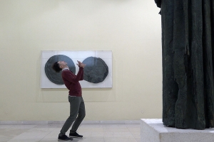 In front of a black monolithic statue, a man in a red sweater stands bent over backward.