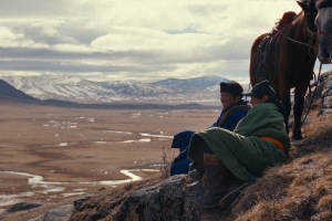 On a steep mountainside, two men sit on the ground next to their horses.