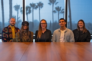 At a conference table, five people in laid back clothing smile at the camera. There are palm trees in the window behind them.