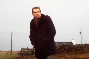 A man in his 30s, wearing a black overcoat, stands on a country road