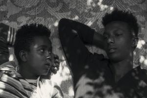 Black and white photo of two young Black men lying on a mat. (l. to r.) Mohammed Arif and Ibrahim Mohammed, two boys from Harar, Ethiopia, as seen in Faya Dayi, directed by Jessica Beshir. Courtesy of Merkhana Films.