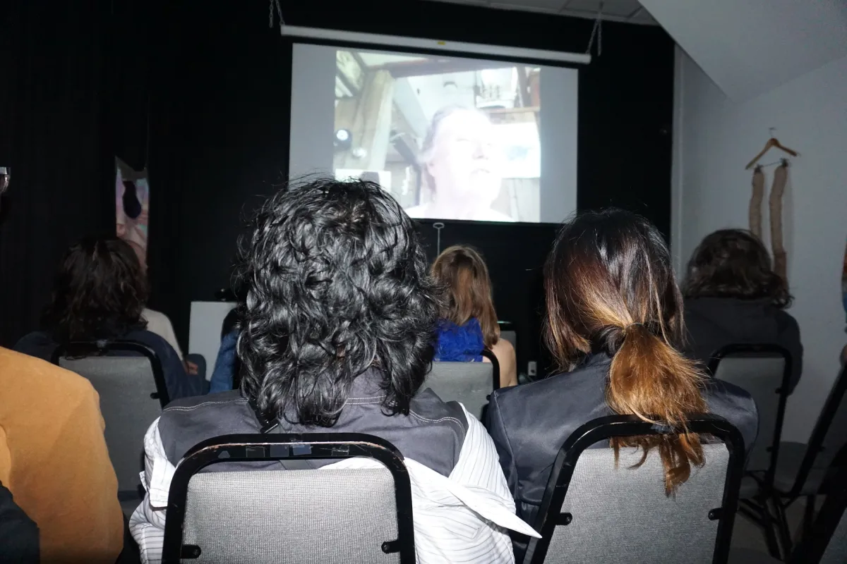 Photograph of the back of an audience, watching a screen hung on a black wall in front of them.