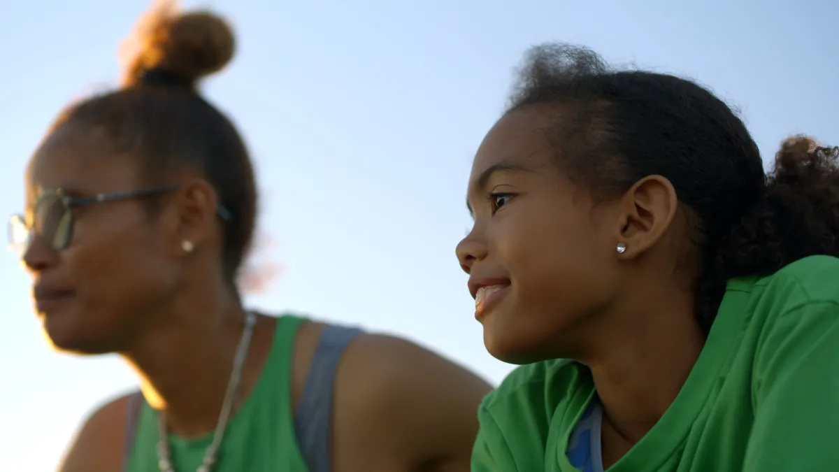 Wearing similarly-colored green shirts, a Black woman and her daughter smile while looking off-screen.
