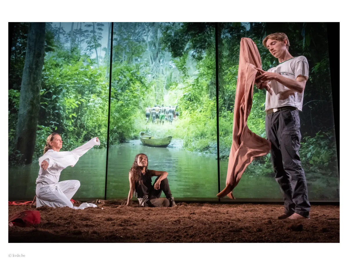 In front of an illuminated screen of the Amazon, three actors sit or stand in the dirt.