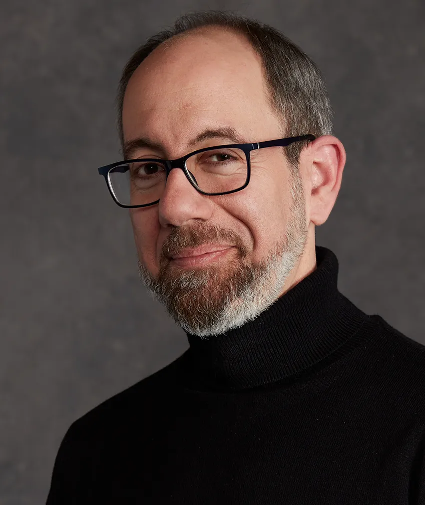 three quarters view posed headshot of a white man in black glasses and a black turtle neck. he is smiling softly and has a brown and gray short trimmed beard. 