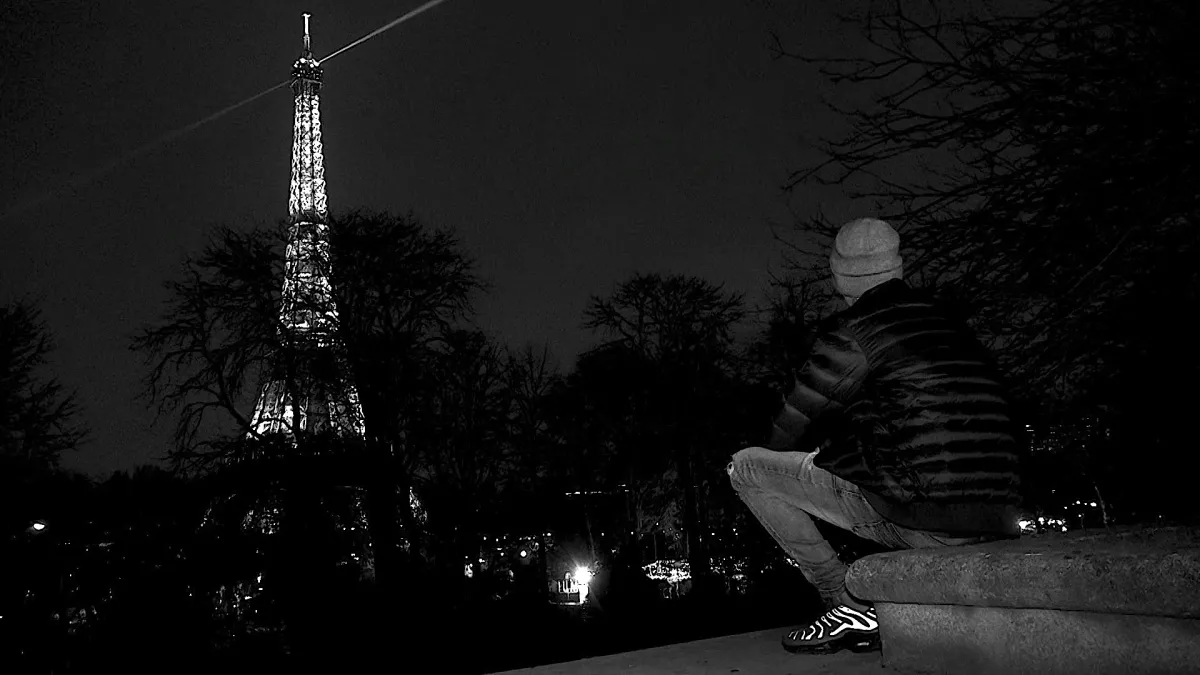 A figure wearing a dark puffy jacket looks at the lit-up Eiffel Tower in the background.