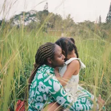 Two black girls, one older and one younger, hug in field of grass.