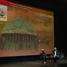 Two men sit at the bottom of a large screen, speaking into microphones.