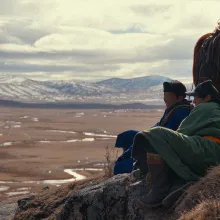 On a steep mountainside, two men sit on the ground next to their horses.